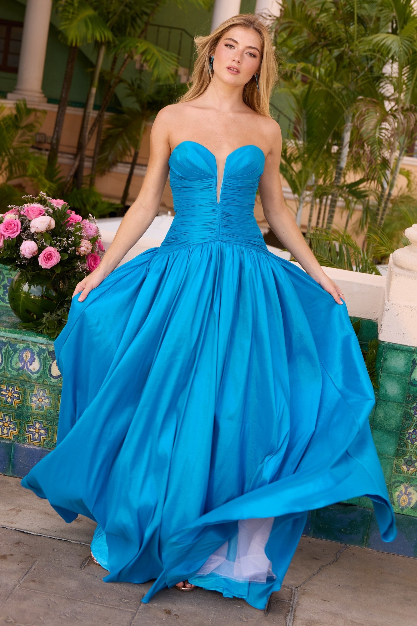 Outdoors, a woman holds the skirt of a vibrant blue Ava Presley 42261 Long Prom Dress with a ruched bodice; pink roses and green foliage bloom in the background.